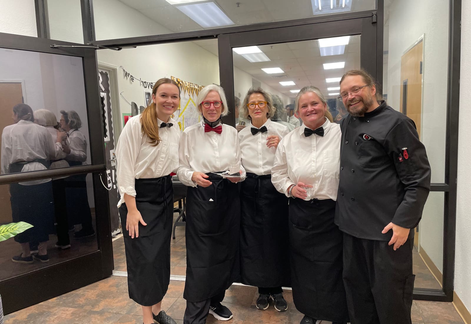 Four servers in white shirts and a chef in a black coat, full length, ready for service.
